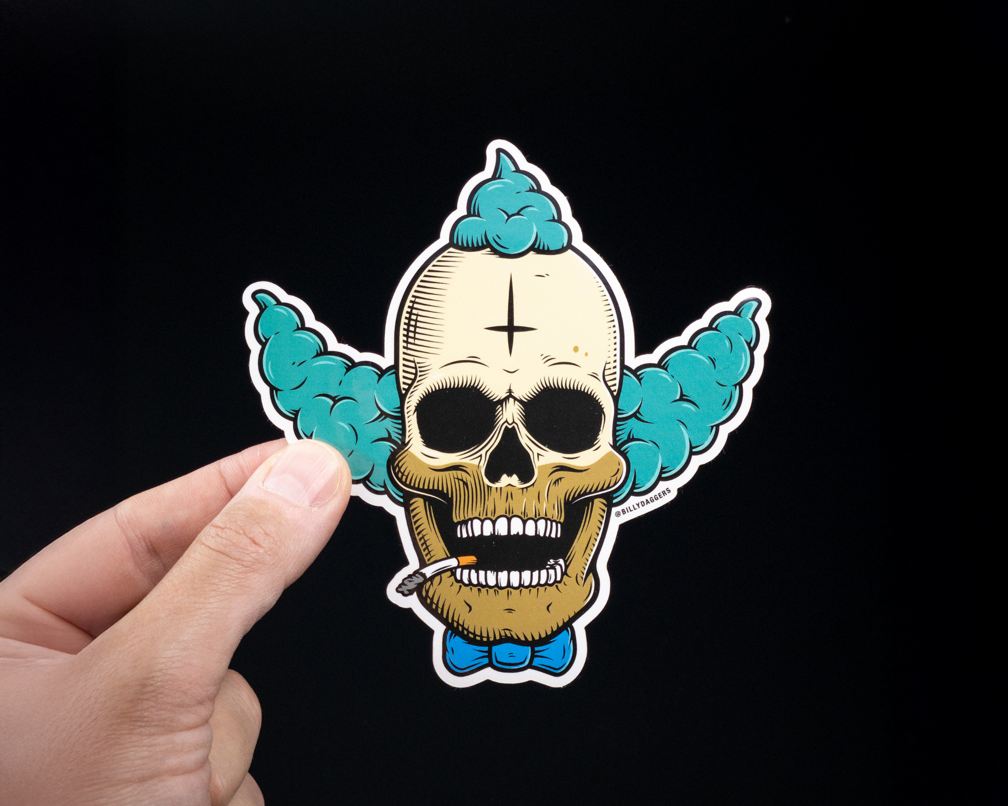 Hand holding a Die cut vinyl sticker of a skull with clown features, including blue hair and a bow tie, against a black background.